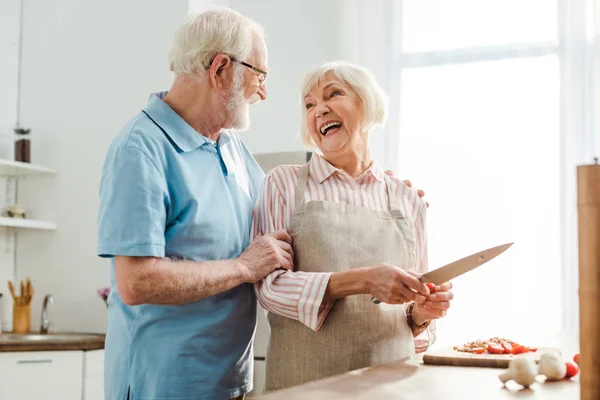 Senior man hugging smiling wife while cutting vegetables on kitchen table — Stock Photo