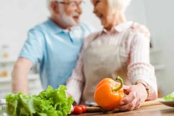 Selective focus of smiling woman looking at husband while holding bell pepper on kitchen table — Stock Photo
