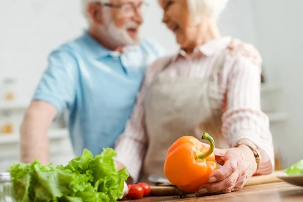 Enfoque selectivo de la mujer sonriente mirando al marido mientras sostiene el pimiento en la mesa de la cocina - foto de stock