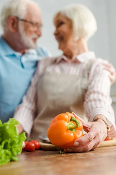 Selektiver Fokus einer Seniorin, die Paprika hält, während sie ihren Mann beim Kochen in der Küche anlächelt — Stockfoto