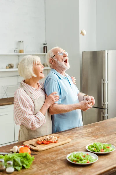 Vista lateral de la mujer mayor riendo mientras el marido vomita seta por verduras maduras en la mesa de la cocina - foto de stock