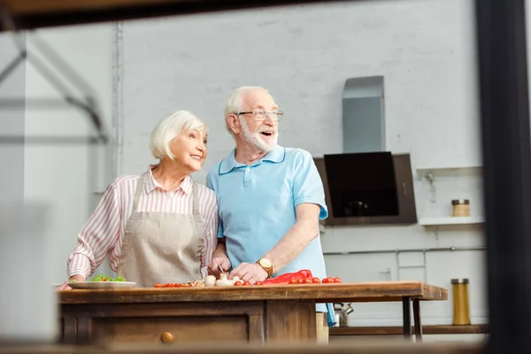 Foco seletivo de casal sênior sorrindo olhando embora enquanto cortando legumes frescos na mesa da cozinha — Fotografia de Stock