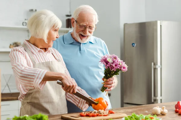 Hombre mayor dando ramo a la esposa sorprendida durante el corte de verduras frescas en la cocina - foto de stock