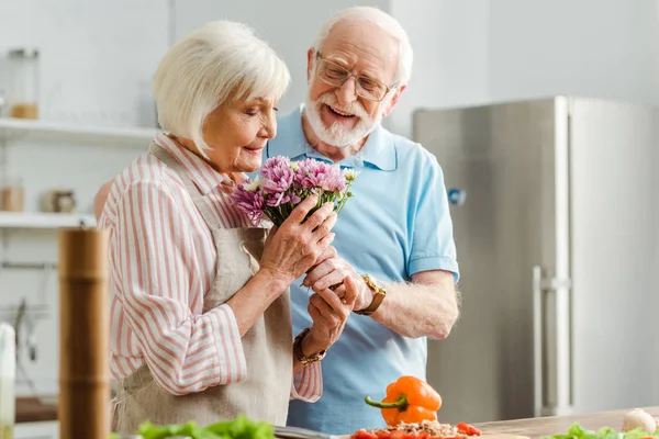 Enfoque selectivo de la mujer mayor aroma ramo al lado sonriente marido y verduras en la mesa de la cocina - foto de stock