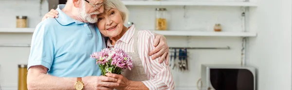 Panoramic shot of senior man hugging smiling wife with bouquet of chrysanthemums in kitchen — Stock Photo