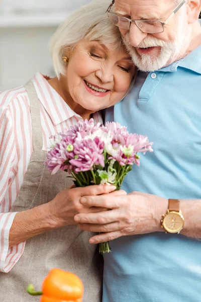 Selective focus of smiling senior couple holding bouquet of chrysanthemums in kitchen — Stock Photo