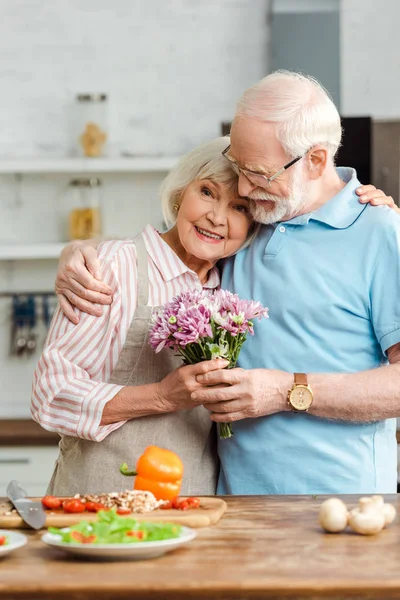 Femme âgée avec bouquet étreignant mari et regardant la caméra à côté des légumes frais sur la table de cuisine — Photo de stock