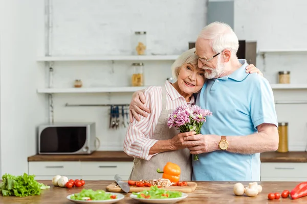 Homme âgé embrassant femme souriante avec bouquet à côté de légumes mûrs sur la table de cuisine — Photo de stock