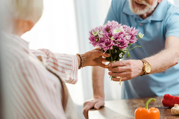 Vista recortada de hombre mayor dando ramo a la esposa al lado de verduras frescas en la mesa de la cocina - foto de stock