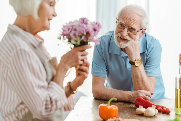 Focus selettivo dell'uomo anziano che guarda la moglie sorridente con bouquet di verdure sul tavolo della cucina — Foto stock