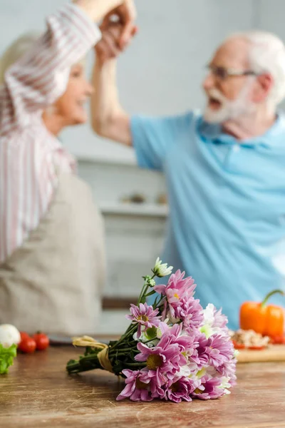 Enfoque selectivo de ramo y verduras en la mesa de la cocina y la pareja de ancianos bailando de fondo - foto de stock