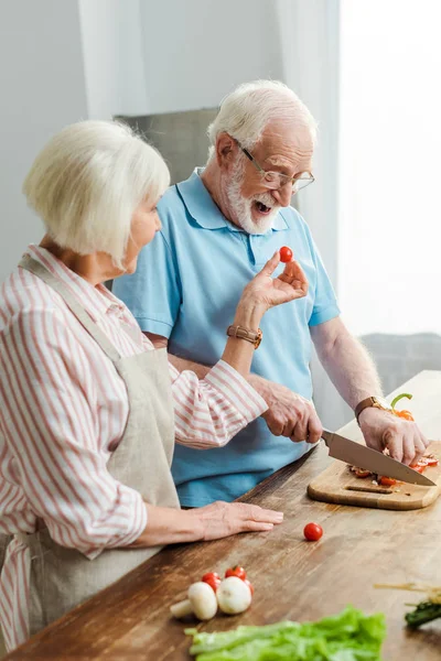 Vista lateral de la mujer mayor dando tomate cherry al marido mientras corta verduras en la mesa de la cocina - foto de stock