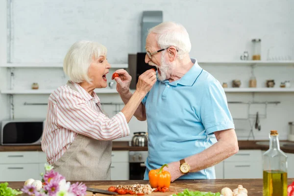 Vista laterale di coppie anziane che si nutrono a vicenda con pomodorini ciliegini mentre cucinano sul tavolo della cucina — Foto stock