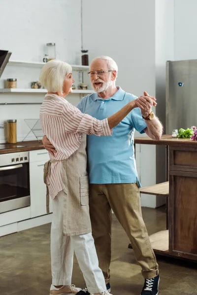 Uomo anziano che sorride alla telecamera mentre balla con la moglie in cucina — Foto stock