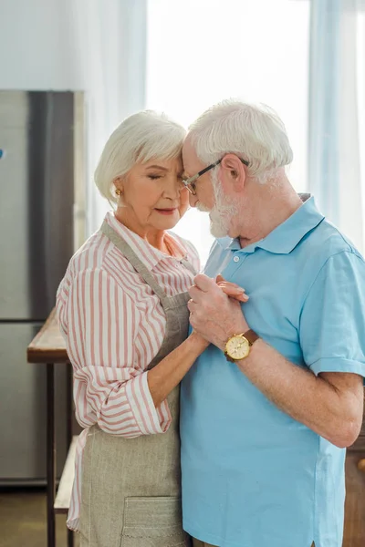 Vista lateral de una pareja de ancianos tomados de la mano mientras se abrazan en la cocina - foto de stock