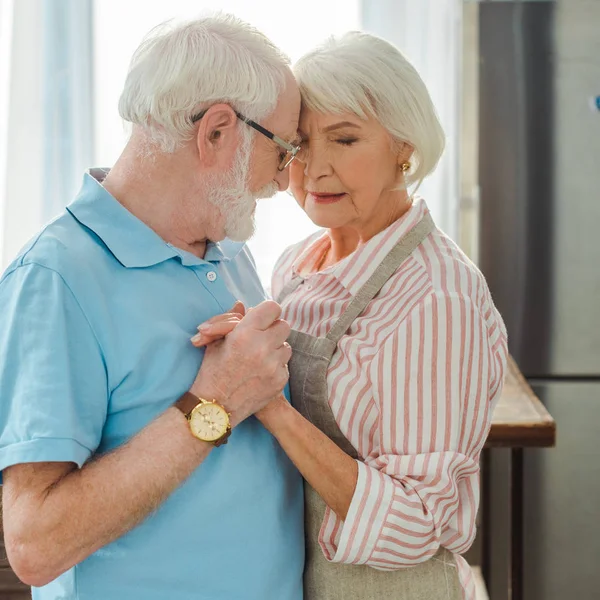 Seitenansicht eines älteren Paares, das sich in der Küche umarmt und Händchen hält — Stockfoto