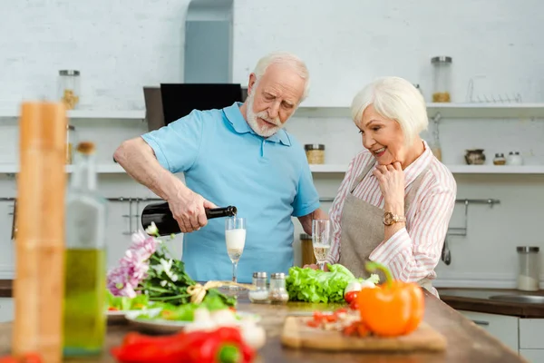 Concentration sélective de la femme souriante regardant mari verser du champagne par bouquet et légumes sur la table de cuisine — Photo de stock