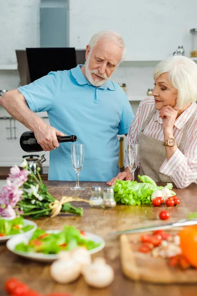 Enfoque selectivo de pareja mayor vertiendo champán por ramo y verduras en la mesa de la cocina - foto de stock