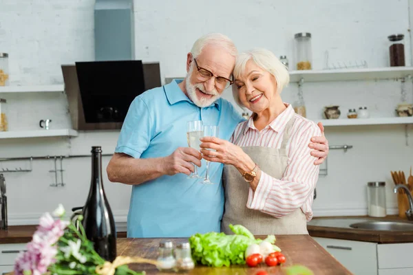 Foco seletivo de sorrindo brindar casal sênior com champanhe por legumes e flores na mesa da cozinha — Fotografia de Stock