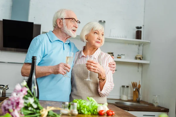 Enfoque selectivo de pareja mayor con copas de champán mirando hacia otro lado junto a ramo y verduras en la mesa de la cocina - foto de stock