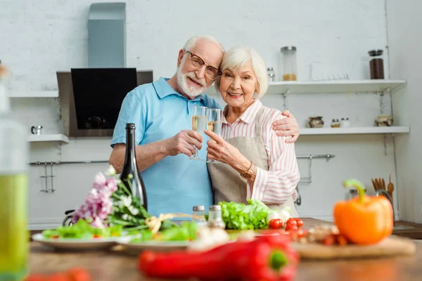 Enfoque selectivo de pareja de ancianos sonriendo a la cámara mientras tintinean con champán por verduras y ramo en la mesa de la cocina — Stock Photo