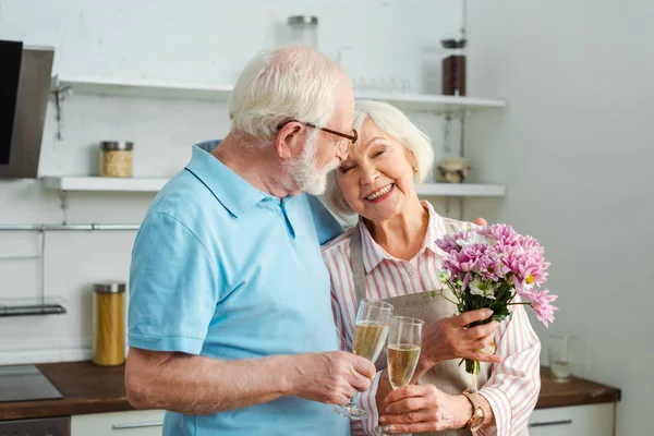 Mulher sênior sorrindo segurando buquê e taça de champanhe com o marido na cozinha — Fotografia de Stock