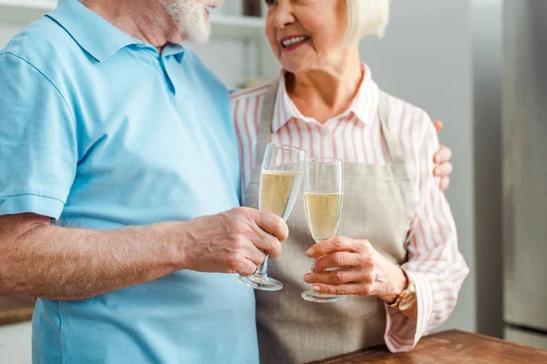 Vista recortada de la pareja de personas mayores sonriéndose entre sí mientras hacen clincing con champán en la cocina - foto de stock