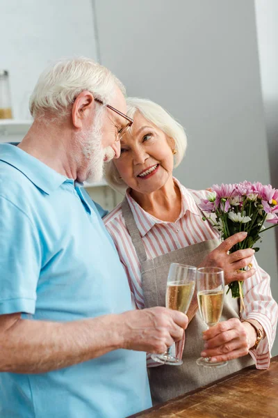 Femme âgée avec bouquet souriant au mari tout en cliquetis avec champagne dans la cuisine — Photo de stock