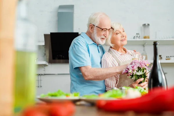 Selective focus of smiling senior couple with bouquet and champagne hugging beside vegetables on kitchen table — Stock Photo