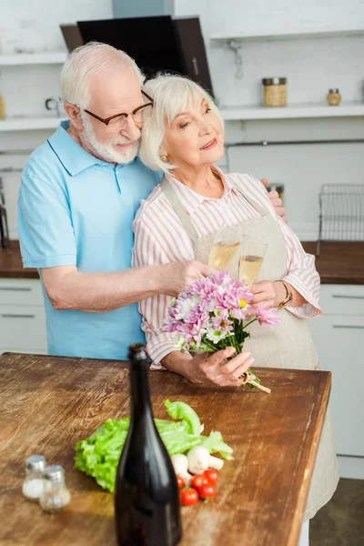 Foco seletivo da mulher idosa segurando buquê e brindando champanhe com o marido na cozinha — Fotografia de Stock