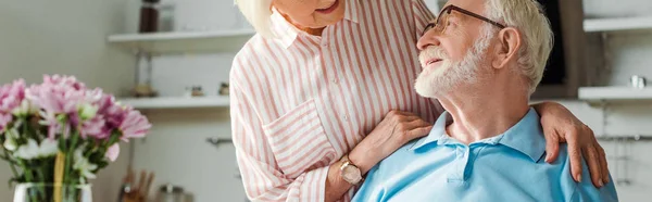 Panoramic shot of smiling woman hugging husband by flowers in vase in kitchen — Stock Photo