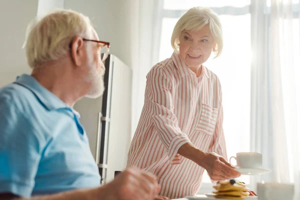 Focus selettivo della donna sorridente che mette caffè e frittelle sul tavolo da marito in cucina — Foto stock