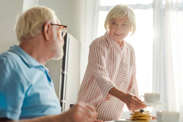 Concentration sélective de la femme souriante mettant du café et des crêpes sur la table par le mari dans la cuisine — Photo de stock