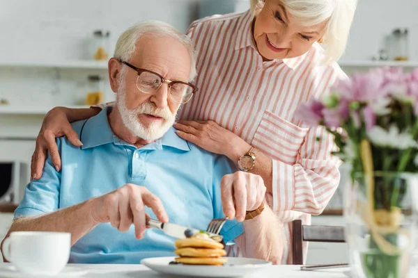 Selective focus of senior man eating pancakes by smiling wife in kitchen — Stock Photo
