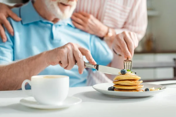 Cropped view of senior man cutting pancakes beside wife in kitchen — Stock Photo