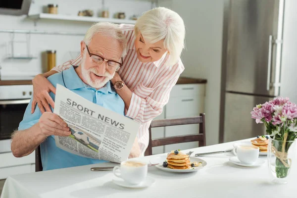 Sonriente pareja de ancianos leyendo el periódico con noticias deportivas por panqueques y café en la mesa - foto de stock