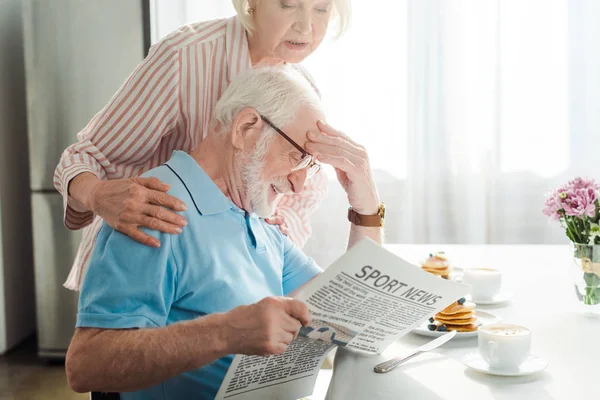 Vista lateral del hombre mayor leyendo el periódico con noticias deportivas por esposa durante el desayuno - foto de stock