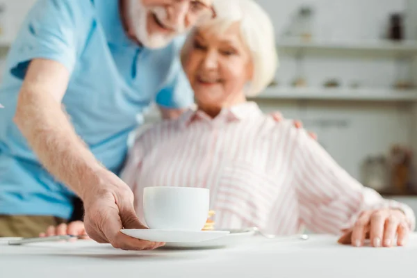 Enfoque selectivo del hombre mayor poniendo taza de café sonriendo esposa en la mesa - foto de stock