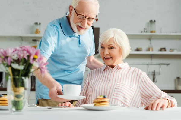 Foco seletivo do homem sênior colocando café na mesa, sorrindo esposa durante o café da manhã — Fotografia de Stock