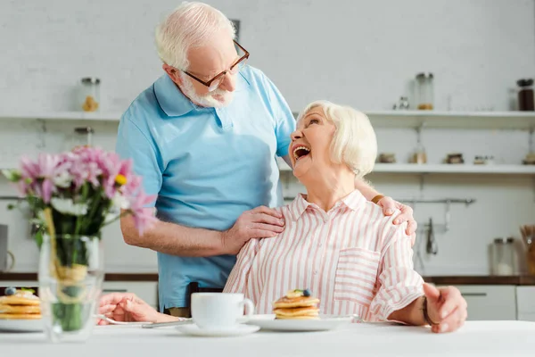 Selektiver Fokus der Seniorin, die lacht, während sie ihren Mann bei Kaffee und Pfannkuchen auf dem Tisch in der Küche anschaut — Stockfoto