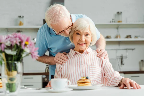 Selective focus of senior man hugging wife looking at camera by pancakes and coffee on table — Stock Photo