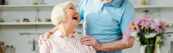 Панорамный снимок пожилой женщины, смеющейся и смотрящей на мужа на кухне — стоковое фото