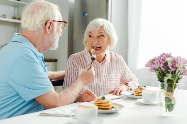 Selective focus of senior man feeding wife with pancake during breakfast in kitchen — Stock Photo