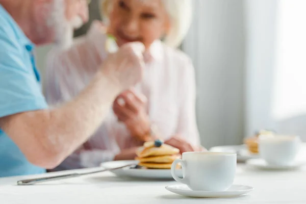Foco seletivo de casal sênior sorrindo um para o outro por café e panquecas na mesa — Fotografia de Stock