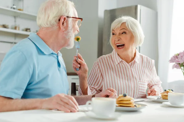 Femme âgée riant tout en nourrissant son mari avec une crêpe pendant le petit déjeuner dans la cuisine — Photo de stock