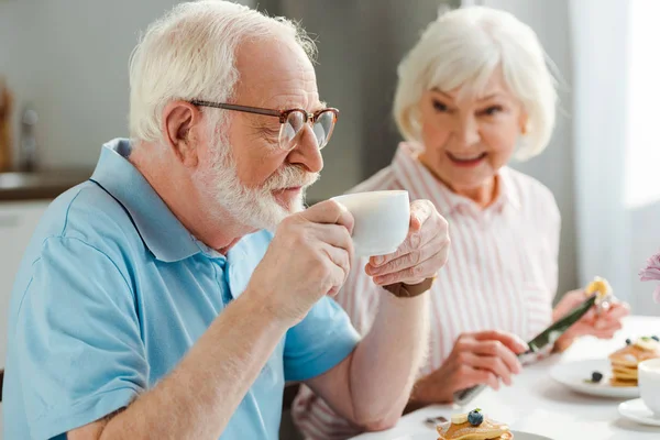 Selektiver Fokus des älteren Mannes beim Kaffeetrinken durch lächelnde Ehefrau mit Pfannkuchen beim Frühstück — Stockfoto