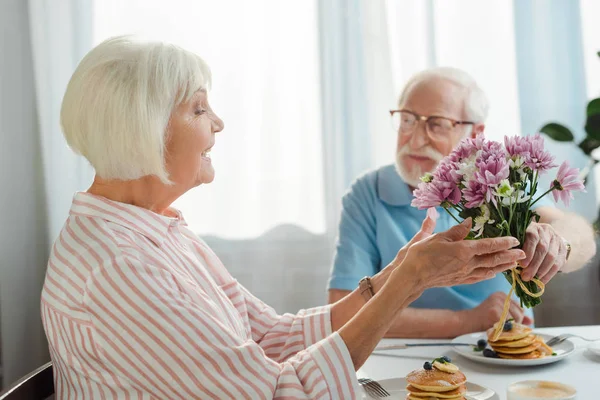 Selective focus of senior man giving bouquet to smiling wife during breakfast in kitchen — Stock Photo