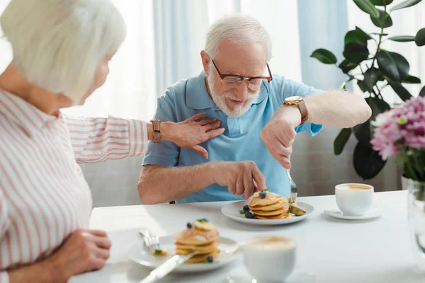 Selective focus of senior woman touching husband with pancakes by coffee and flowers on table — Stock Photo