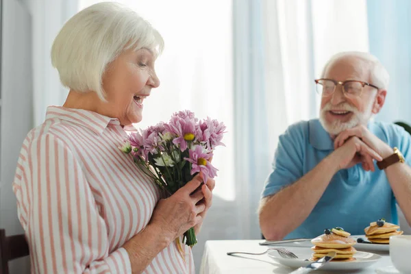 Concentration sélective de la femme tenant bouquet en souriant mari à table avec café et crêpes — Photo de stock