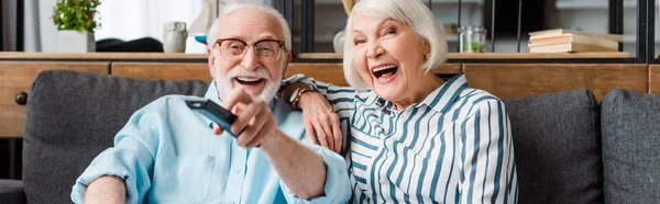 Panoramaaufnahme eines älteren Ehepaares, das zu Hause auf dem Sofa vor dem Fernseher lacht — Stockfoto